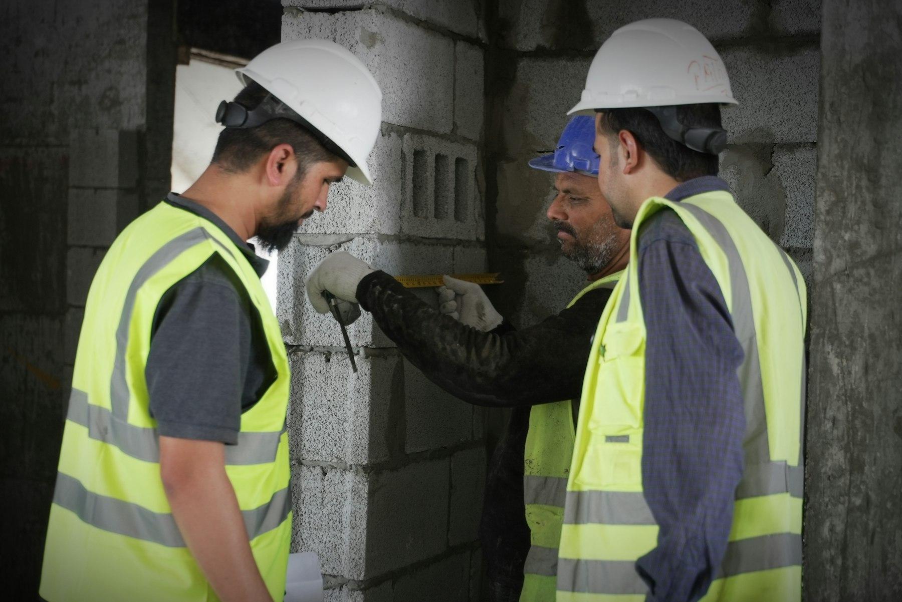 Two site workers in high-visibility vests checking a block wall